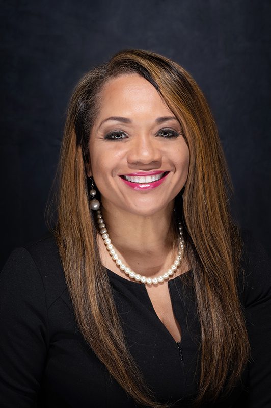 A woman with long hair and pearls in front of a black background.