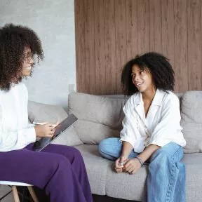 Two women sitting on a couch talking to each other.
