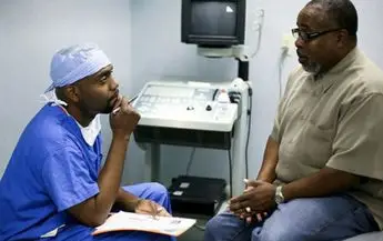 A man in blue scrubs and glasses talking to another man.