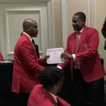 Two men in red jackets are holding a paper.